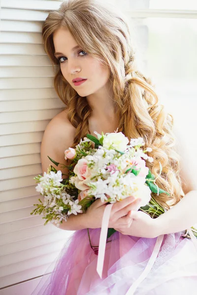 Ritratto di bella giovane ragazza in un vestito dolce e lunghi capelli biondi seduta su una finestra con composizione di fiori — Foto Stock