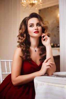 Close-up portrait of beautiful young woman in gorgeous red velvet evening dress sitting by the table in expensive interior