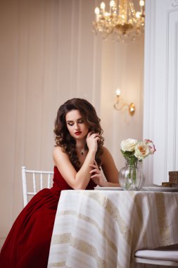 Close-up portrait of beautiful young woman in gorgeous red velvet evening dress sitting by the table in expensive interior