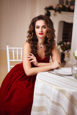 Close-up portrait of beautiful young woman in gorgeous red velvet evening dress sitting by the table in expensive interior