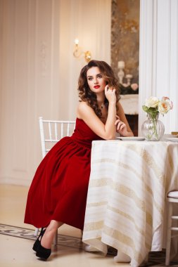 Close-up portrait of beautiful young woman in gorgeous red velvet evening dress sitting by the table in expensive interior