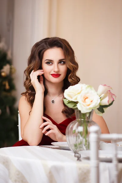 Close-up portrait of beautiful young woman in gorgeous red velvet evening dress sitting by the table in expensive interior — Zdjęcie stockowe