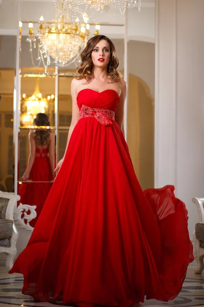 美しい若い女性の完璧なメイクアップと高価な高級インテリアでゴージャスな赤いイブニング ドレスのヘアー スタイル — ストック写真