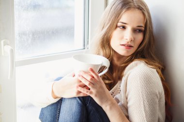 Portrait of young lady wearing white pullover sitting on a windowsill