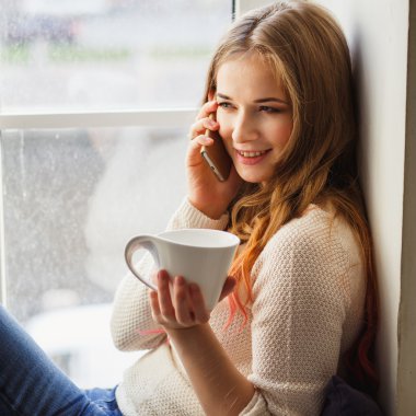 Portrait of young lady wearing white pullover sitting on a windowsill and talking by phone