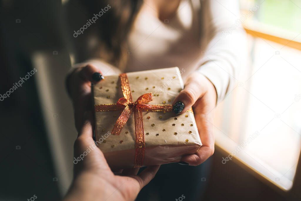 Young women with black glitter nails giving a Christmas present box to his friend. Hands hold new year gift box. decorated with craft paper, red and golden ribbon and stars. Family gift and holiday concept.