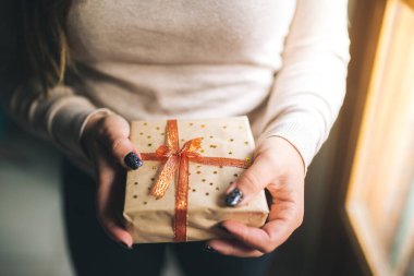 Parlak tırnakları olan genç kadınlar Noel hediyesi kutusunu tutuyorlar ve kamerada gösteriyorlar. Eller yeni yıl hediye kutusunu tutar. Zanaat kağıdı, kırmızı ve altın kurdele ve yıldızlarla süslenmiş. Aile hediyesi konsepti.