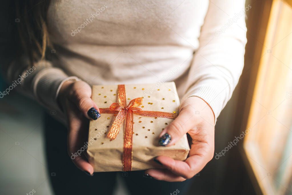 Young women with glitter nails holding Christmas present box and showing it in camera. Hands hold new year gift box. decorated with craft paper, red and golden ribbon and stars. Family gift concept.