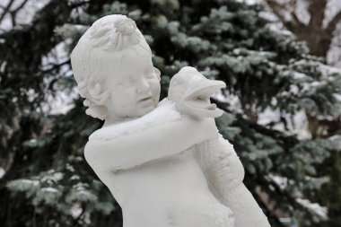 Kaz tutan bir çocuğun klasik beyaz heykeli, hafif bir kar tabakasıyla kaplı, kış bahçesindeki yeşil ağaçların arasında duruyor..