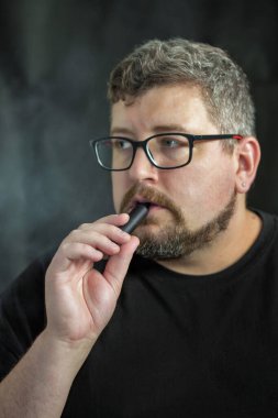 Elektronik sigara içen, gözlük ve küpe takan, kızıl saçlı, sakallı ve siyah arka planda bıyıklı beyaz bir adamın stüdyo portresi..