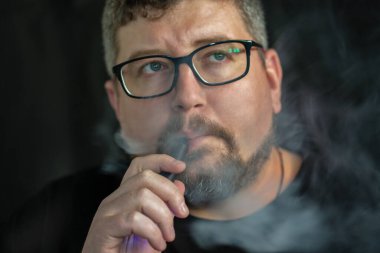 Elektronik sigara içen, gözlük ve küpe takan, kızıl saçlı, sakallı ve siyah arka planda bıyıklı beyaz bir adamın stüdyo portresi..