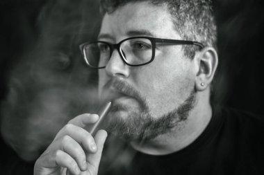 Elektronik sigara içen, siyah arka planda sakal ve bıyıklı, gözlük ve küpe takan beyaz bir adamın stüdyo portresi..