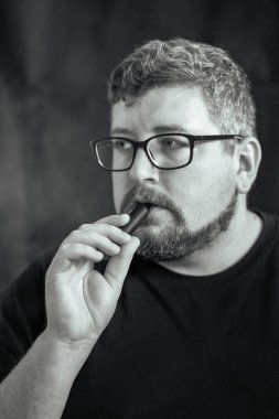 Elektronik sigara içen, gözlük ve küpe takan, kızıl saçlı, sakallı ve siyah arka planda bıyıklı beyaz bir adamın stüdyo portresi..