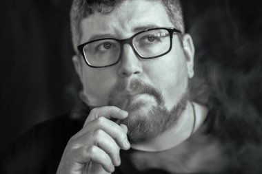 Elektronik sigara içen, gözlük ve küpe takan, kızıl saçlı, sakallı ve siyah arka planda bıyıklı beyaz bir adamın stüdyo portresi..