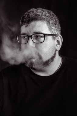 Elektronik sigara içen, siyah arka planda sakal ve bıyıklı, gözlük ve küpe takan beyaz bir adamın stüdyo portresi..