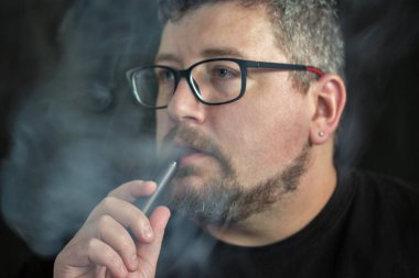 Elektronik sigara içen, gözlük ve küpe takan, kızıl saçlı, sakallı ve siyah arka planda bıyıklı beyaz bir adamın stüdyo portresi..