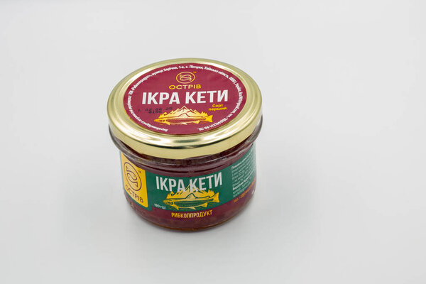Kyiv, Ukraine - March 7, 2025: Ostrov brand red salmon caviar in glass jar with gold lid. Ukrainian fish product labeled as premium grade caviar packaging displayed closeup on white surface.