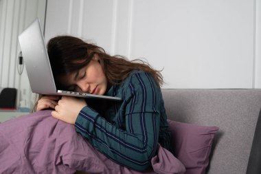 Yataktaki genç kadın dizüstü bilgisayarda uyuyakalmış. Sabah oldu ama şimdiden yoruldu.