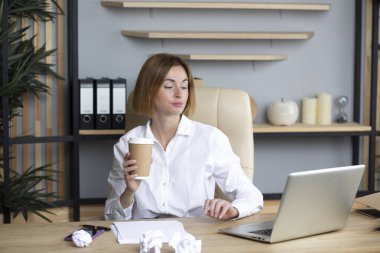 Kendine güvenen iş kadını laptopta çalışıyor ve modern ofiste elinde kahve fincanı tutuyor.