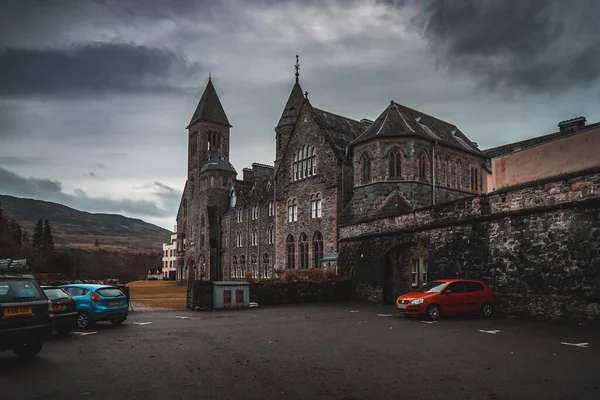 FORT AUGUSTUS, SCOTLAND, DECEMBER 17, 2018: The Abbey Highland Club, full of mold and lichen in its stone walls, under heavy cloudscape with mountains in the background, aside Loch Ness. — Stock Photo, Image