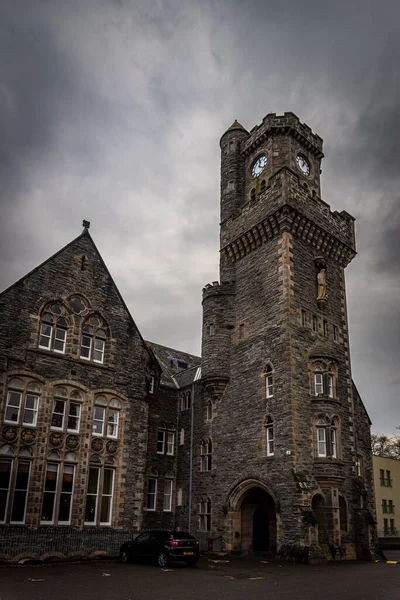 FORT AUGUSTUS, SCOTLAND, DECEMBER 17, 2018: The Abbey Highland Club clock tower, full of mold and lichen in its stone walls, under heavy cloudscape with mountains in the background, aside Loch Ness. — Stock Photo, Image