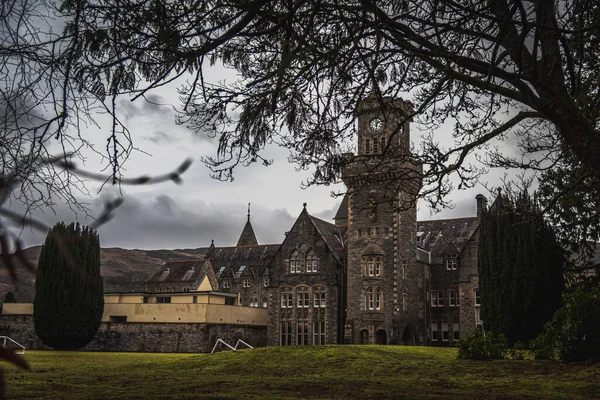 FORT AUGUSTUS, SCOTLAND, DECEMBER 17, 2018: The Abbey Highland Club clock tower, full of mold and lichen in its stone walls, under heavy cloudscape with mountains in the background, aside Loch Ness. — Stock Photo, Image