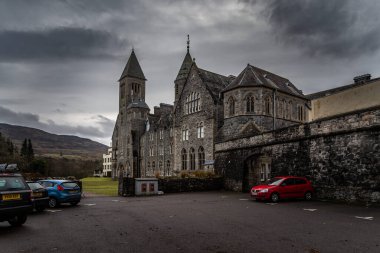 Fort AUGUSTUS, SCOTLAND, 17 ARALIK 2018: Taştan duvarlarında küf ve liken dolu Abbey Highland Kulübü, arka planda dağlarla çevrili yoğun bulutlar altında,.