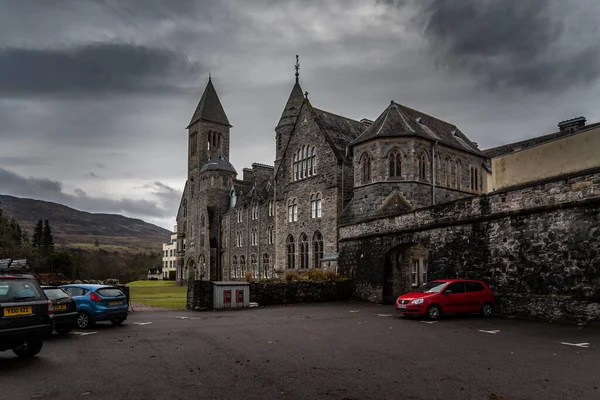 FORT AUGUSTUS, SCOTLAND, DECEMBER 17, 2018: The Abbey Highland Club, full of mold and lichen in its stone walls, under heavy cloudscape with mountains in the background, aside Loch Ness. — Stock Photo, Image