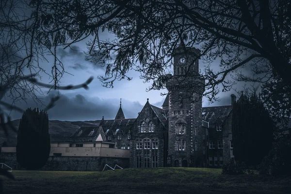 FORT AUGUSTUS, SCOTLAND, DECEMBER 17, 2018: The Abbey Highland Club clock tower, full of mold and lichen in its stone walls, under heavy cloudscape with mountains in the background, aside Loch Ness. — Stock Photo, Image