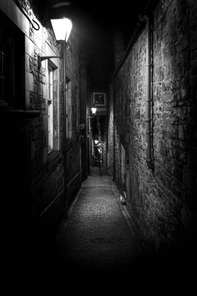 A dark creepy narrow European alley at night, surrounded by bricks and cobblestone. Illuminated only with some street lamps. Concept of scared or being alone and frightened.