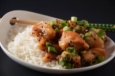 Baharatlı teriyaki tavuğu pirinçli fileto, yeşil soğan ve siyah arkaplanda beyaz tabakta siyah susam tohumları.