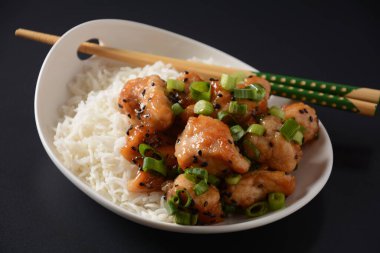 Baharatlı teriyaki tavuğu pirinçli fileto, yeşil soğan ve siyah arkaplanda beyaz tabakta siyah susam tohumları.