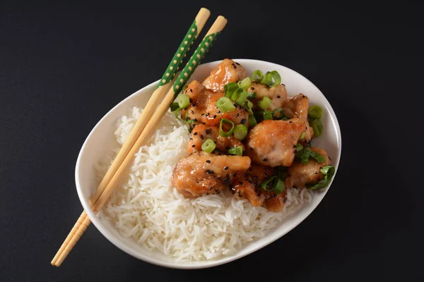 Spicy teriyaki chicken fillet pieces with rice, green onions and black sesame seeds on white plate on a black background