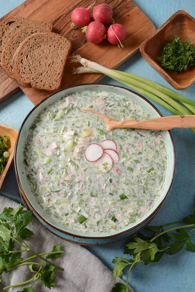 Cold soup okroshka with sausage, potato, egg, radish, cucumber, greens and kvass in a bowl, bread on wicker napkin. Traditional Russian summer cold soup with kvass