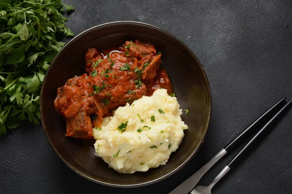 Soslu biftek parçaları, patates püresi ve siyah arka planda otlar.