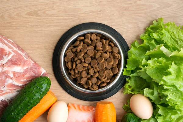 Dry pet dog food with natural ingredients. Raw meat, fish, vegetables, eggs and salad near bowl with dry pet feed on wooden background. concept of correct balanced nutrition for pet, flatly