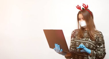Noel alışverişi kavramı, salgın için tebrikler. Tıbbi maskeli genç bir kadın, eldivenler ve dizüstü bilgisayarlı geleneksel kırmızı geyik boynuzları. kopyalama alanı ile