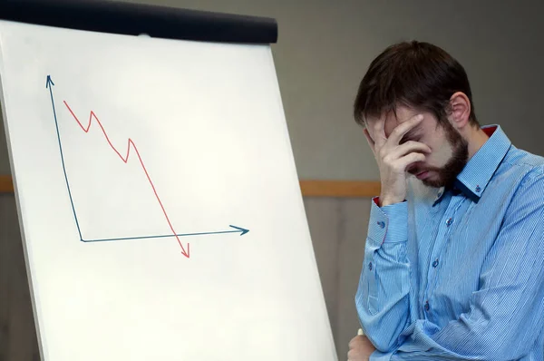 Frustrated business man standing near white board in office, covers his face with his hand at the falling graph of a stock market struck in financial crisis