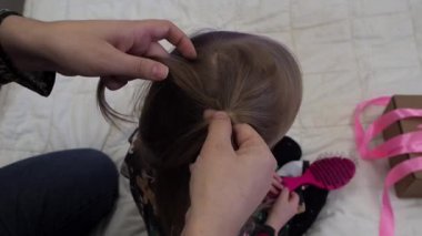 Anne, sarı saçlı kızın saçını örüyor. Kafaya örgü örme işlemi yapılıyor. Eller yukarı..