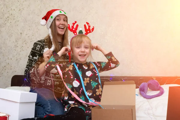 woman and kid girl packaging Christmas gifts for friends and family. mom wearing red Santa Claus hat with child in deer antlers have fun, play, preparation for xmas holidays. cozy celebration concept.