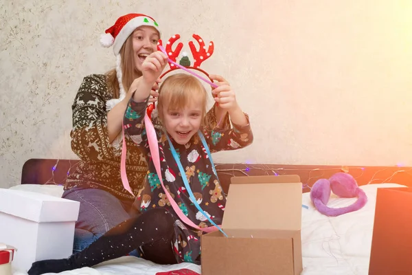 女性と子供の女の子は友人や家族のためのクリスマスプレゼントを包装します。鹿の角の子供と赤いサンタクロースの帽子を身に着けているお母さんは楽しみ、遊び、クリスマス休暇とお祝いの準備をしています. — ストック写真