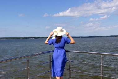 Hasır şapkalı genç kadın zarif elbiseler içindeki iskelede kalıp manzaranın tadını çıkarıyor..