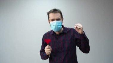 Tıbbi maske takan, kırmızı kalp takan ve dans eden güzel bir adamın portresi. İnternette sevgililer gününü kutlayan adamdan tebrikler. Romantik.