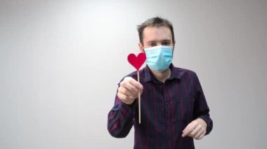 Tıbbi maske takan, kırmızı kalp takan ve dans eden güzel bir adamın portresi. İnternette sevgililer gününü kutlayan adamdan tebrikler. Romantik.