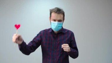 Tıbbi maskeli, kırmızı kalp takan ve dans eden güzel bir adam portresi. İnternette sevgililer gününü kutlayan adamdan tebrikler. Romantik.