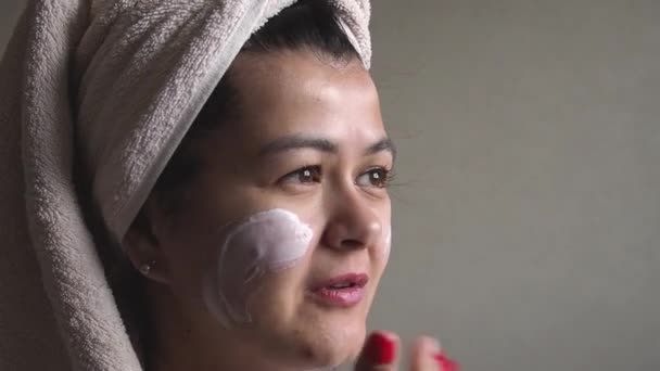 Porträtt av en kvinna som applicerar en kosmetisk kräm på ansiktet med handduk på huvudet. Skönhetsbehandlingar och spa hemma. — Stockvideo