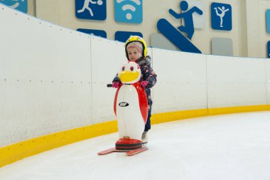 3 yaşındaki kız, kapalı bir buz pateni pistinde paten kaymayı öğreniyor. Plastik bir penguen, patenci kız tarafından destek için itiliyor. Küçük çocuklar için eğlenceli kış aktivitesi