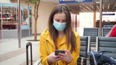 Bekleme odasında tıbbi maske koruması olan beyaz kadın turist akıllı telefondan çalışıyor. Havaalanı istasyonunda oturan sarı elbiseli gezgin kadın. Gezginler için yeni normal.