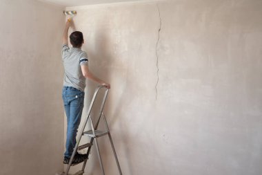 Duvarı silindirle kaplayan bir adam. İçinin fotokopi alanıyla onarılması. Genç erkek dekoratör boş odada bir duvar boyuyor. Yenileme işini kendin yap.