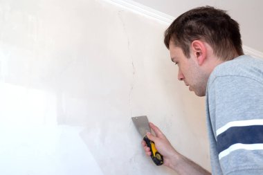 İnşaat işçisi bir evin çatlak duvarını onarıyor, duvara çimento yapıştırıyor. İnşaatçı, macun bıçağıyla duvardaki bir çatlağa beyaz çimento sürüyor. 